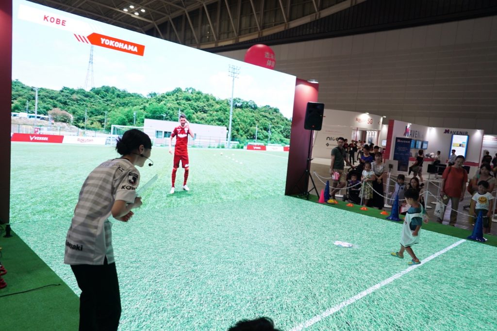 A young footballer passes the ball to Vissel Kobe star David Villa at a customized augmented reality booth.